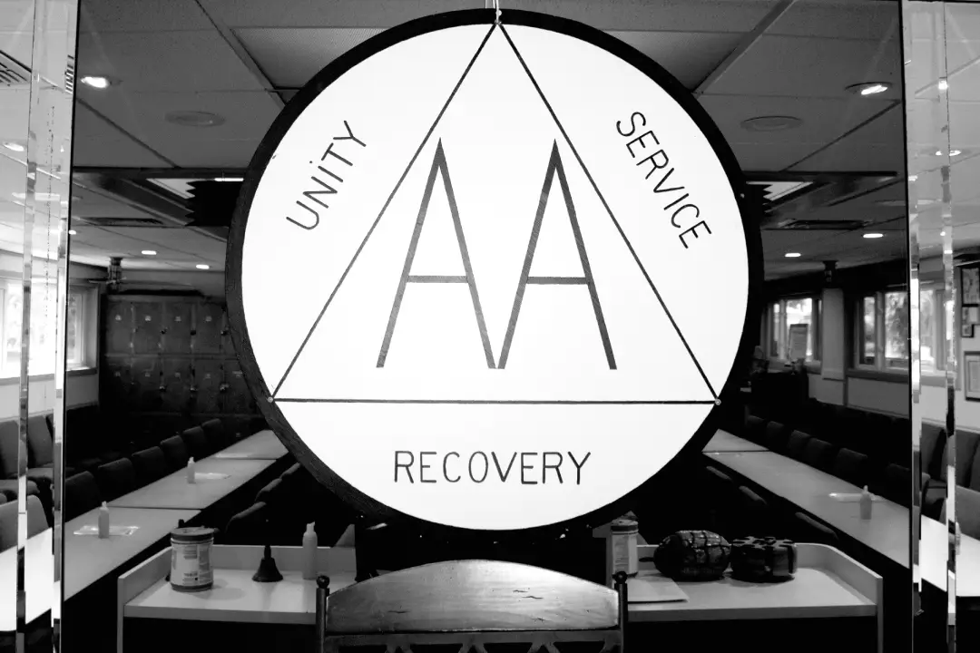 Alcoholics Anonymous symbol featuring _Unity, Service, Recovery_ in a meeting room with chairs, tables, and an American flag visible